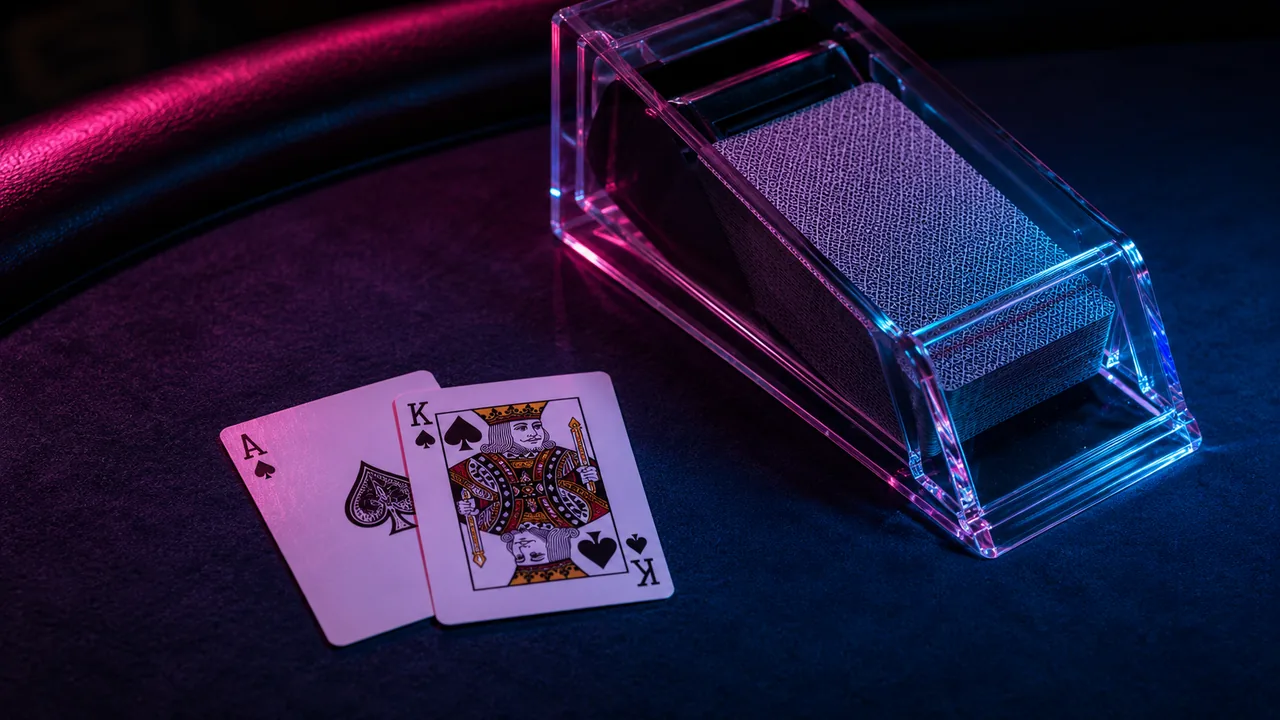 An ace and a king on dark casino felt beside a transparent card shoe, lit by neon.