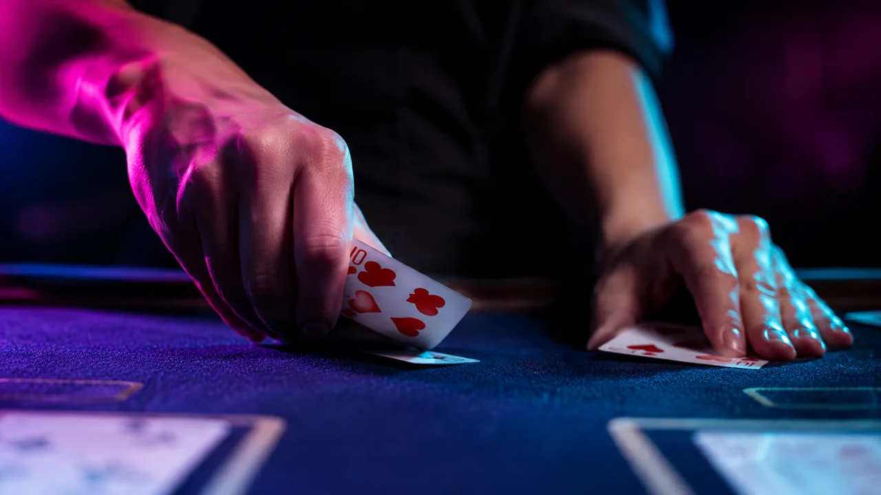 A dealer's hands dealing playing cards across blue casino felt under neon light.